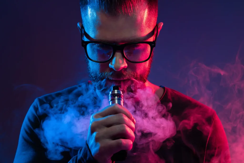 A guy with glasses vaping while smoke coming out of his mouth.
